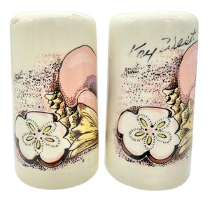 May include: A pair of ceramic salt and pepper shakers with a cream-colored base. Each shaker features a floral design with pink, yellow, and brown accents. The text "Key West" is written on one shaker.