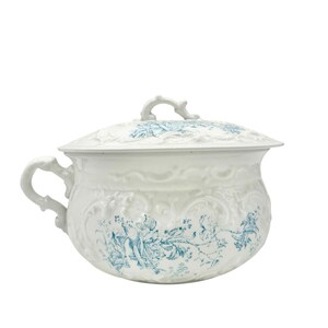 May include: A white ceramic chamber pot with a matching lid, featuring an ornate embossed scrollwork pattern and a blue floral transfer print. The vessel includes a single side handle and a decorative finial on the lid, showcasing a classic design.