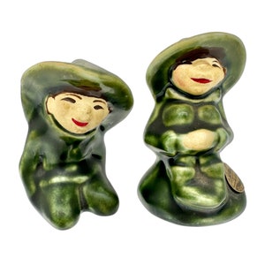 May include: A pair of green ceramic salt and pepper shakers shaped like people wearing hats. The shakers have painted faces with smiles and are sitting on a white background.