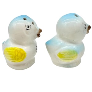 Pode incluir: Um par de saleiros e pimenteiros de cerâmica em forma de pássaros de desenho animado. Os pássaros são brancos com detalhes azuis na cabeça e nas asas, e manchas amarelas nas laterais. Eles têm bicos marrons e olhos pretos.