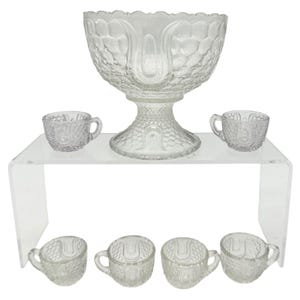 Op de afbeelding: Een punchbowlset van helder glas bestaande uit een grote schaal op voet en zes bijpassende kleine kopjes. Elk stuk is gemaakt van geperst glas met een gestructureerd, herhalend schubpatroon en gladde, boogvormige verticale panelen.