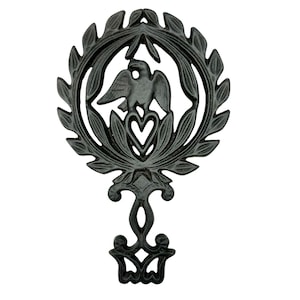 May include: Black cast iron trivet with an eagle and heart design. The trivet features a decorative oval frame with a handle and a detailed floral and leaf pattern. The eagle is centered within the frame.