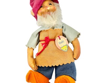 May include: A vintage gnome doll with a red pointed hat, white beard, and orange felt shoes. The doll wears a light blue shirt, a burlap apron tied with a red string, and blue denim pants. A tag reads "Gucki" with a yellow bear.