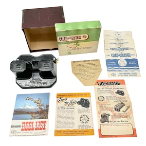 May include: A vintage View-Master set with a black stereoscope viewer, boxes, and various reels and brochures. The green and white box reads "View-Master Stereoscope." The set includes a reel list and instructions. The items are arranged on a white surface.
