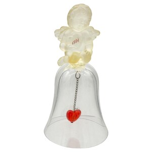 May include: Transparent bell-shaped ornament with a cherub figurine on top. The cherub is light yellow and has wings. The year "1991" is printed in red on the cherub's chest. A red heart hangs from a chain inside the bell.
