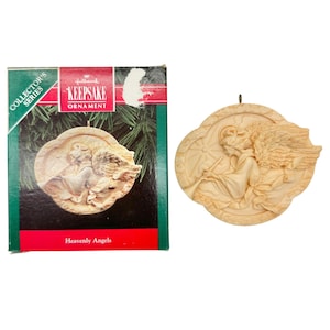 May include: A cream-colored, oval-shaped Hallmark Keepsake Ornament featuring a relief of an angel playing a trumpet. The ornament is accompanied by its original box, which displays the text Collector's Series, Hallmark Keepsake Ornament, and Heavenly Angels.