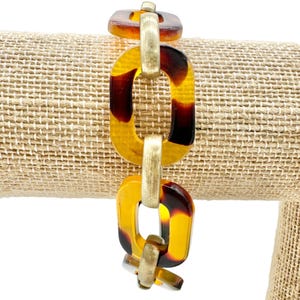 May include: A chain link bracelet featuring alternating rectangular links made of translucent amber and dark brown tortoiseshell patterned resin and brushed gold-tone metal. The links are connected in a repeating pattern to form the bracelet structure.