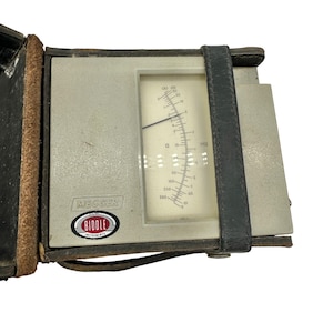 Könnte beinhalten: Ein Vintage-Elektroprüfgerät von Megger Biddle Instruments. Das Gerät hat eine graue Oberfläche mit einer Nadelanzeige und einem schwarzen Lederriemen. Das Messgerät zeigt Messungen in Ohm und Megaohm an. Das Gerät befindet sich in einem schwarzen Lederetui.