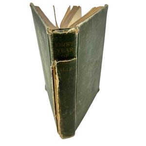 May include: A vintage book with a green cover and the title "First Year of Allen" on the spine. The book's pages are aged, and the binding shows signs of wear, indicating its age and use over time.