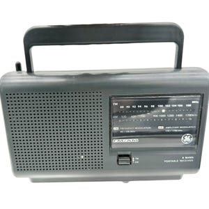 Puede incluir: Una radio portátil gris oscuro con un asa negra. La radio tiene una rejilla de altavoz, un dial de sintonización y el texto "FM/AM" y "2 Band Portable Receiver". La radio tiene el logotipo de GE.