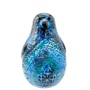 May include: A ceramic bird figurine featuring a glossy glaze in shades of deep blue, turquoise, and teal. The surface is decorated with embossed geometric patterns, including circular motifs and a central star-like design on the front.