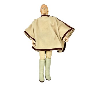 Op de afbeelding: Vintage actiefiguur van een personage uit een sciencefictionfranchise. De figuur draagt een crèmekleurige poncho met donkerrode rand, crèmekleurige laarzen en een beige broek. De figuur heeft blond haar en poseert tegen een witte achtergrond.