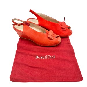 Op de afbeelding: Een paar felrode slingback-wigheels met open teen en strikaccenten. De schoenen worden gepresenteerd op een bijpassende rode stoffen tas met de witte tekst "BeautiFeel".