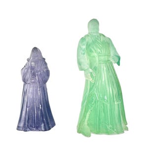 May include: Two translucent figures of robed characters. One is a smaller, hooded figure in a lavender hue. The other is a larger figure in a light green color, with a detailed robe and a standing pose. Both figures are set against a white background.