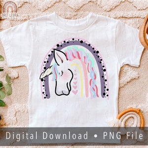 May include: A white t-shirt with a rainbow and unicorn design. The rainbow is made up of pink, purple, blue, green, and yellow stripes. The unicorn is white with a pink mane and tail. The design is surrounded by black dots.