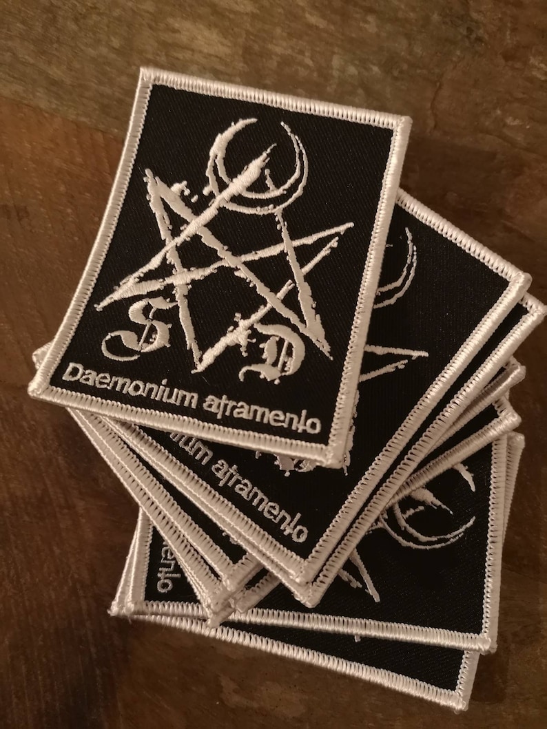 Peut inclure: Une pile de patchs en tissu noir avec des motifs brod&eacute;s blancs. Les patchs pr&eacute;sentent un pentagramme entour&eacute; d'un cercle et le texte "daemonium atramento".