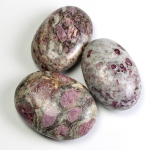 May include: Three oval stones in shades of grey, brown, and pink. The stones have a speckled appearance, with larger pink crystal formations. The stones are smooth and polished, with a natural, organic aesthetic.