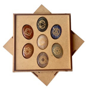 May include: Set of seven chakra stones in a wooden box. Each stone is a different color and has a gold symbol on it. The stones are arranged in a circle, with the root chakra stone at the bottom and the crown chakra stone at the top.