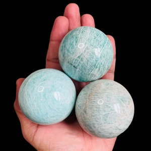 May include: Three smooth, green, polished amazonite spheres, each with a unique pattern of white veins, are held in a hand.
