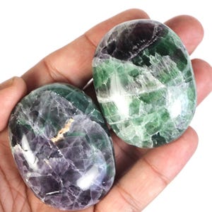 May include: Two green, purple and white fluorite palm stones. The stones are smooth and polished, with a variety of patterns and textures.