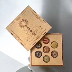 May include: A wooden box with a lid that opens to reveal seven round stones with gold symbols. The lid has a laser-engraved image of a person in a yoga pose with seven chakras.