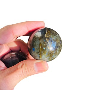 May include: Two polished labradorite spheres, one held in a hand, with iridescent blue and green hues.