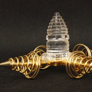 May include: A clear crystal pyramid sits atop a gold metal stand with three spiral arms. The stand has a pointed tip on each arm.