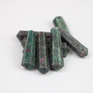 May include: Several green and red ruby zoisite crystal wands. The hexagonal wands are of varying sizes. The crystals have a speckled appearance with green and red colours. The crystals are arranged on a white surface.