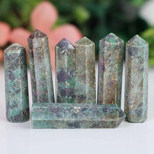 May include: Seven green, purple, and brown gemstone points, also known as crystal wands, are arranged in a row on a white surface.