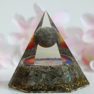 May include: A clear crystal pyramid with a gray sphere inside. The pyramid has a colorful base with a sunset design and a layer of gold glitter.