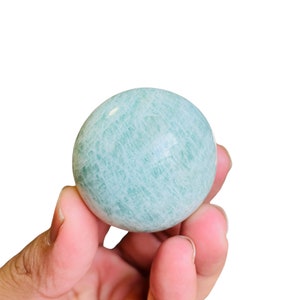 May include: A smooth, green and white, polished Amazonite sphere. The sphere is about the size of a golf ball and is being held in a hand.