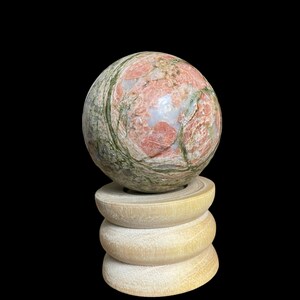 May include: A green and pink sphere made of polished stone sits on a wooden stand. The sphere has a swirling pattern of green and pink.
