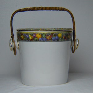 Puede incluir: Cubo de hielo de porcelana blanca con un motivo de frutas y un asa de mimbre marrón. La tapa tiene un borde dorado.