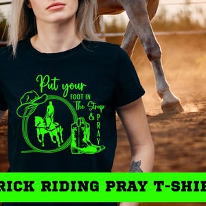 May include: Black t-shirt with a neon green graphic featuring a cowboy hat, a person riding a horse, and a cowboy boot. Text on the shirt reads "Put your foot in the strap & pray." The words "TRICK RIDING PRAY T-SHIRT" are at the bottom.