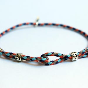 Puede incluir: Una pulsera trenzada con un diseño colorido. La pulsera presenta un patrón de hilos naranja, azul y gris. Cuentas de metal plateado se colocan a intervalos a lo largo de la pulsera. Sobre un fondo blanco.