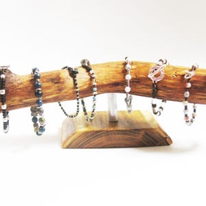 Op de afbeelding: Een houten tak met een natuurlijke bastafwerking, gemonteerd op een houten voet, wordt gebruikt als displaystandaard voor verschillende kralenarmbanden. De armbanden zijn gemaakt met kralen in verschillende kleuren, waaronder zwart, wit, blauw en bruin.