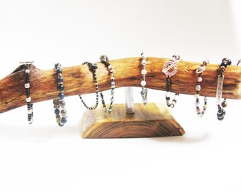 Könnte beinhalten: Ein Ast aus Holz mit natürlicher Rindenoptik, auf einem Holzsockel montiert, dient als Präsentationsständer für verschiedene Perlenarmbänder. Die Armbänder sind aus Perlen in verschiedenen Farben gefertigt, darunter Schwarz, Weiß, Blau und Braun.