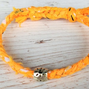 Può includere: Un braccialetto in tessuto intrecciato giallo con un piccolo ciondolo dorato a forma di fiore.