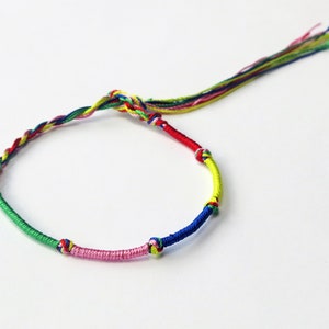 Op de afbeelding: Een veelkleurig gevlochten vriendschapsarmband met een roze, groene, blauwe, gele en rode draad. De armband heeft een lange kwast.