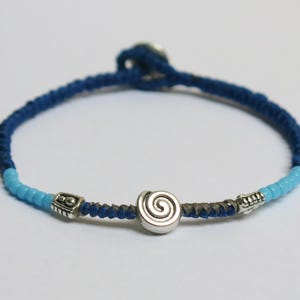 Può includere: Braccialetto blu fatto a mano con un ciondolo a spirale argentato. Il braccialetto presenta un design intrecciato con perline azzurre e dettagli argentati. Il braccialetto ha una circonferenza di circa 18 cm.