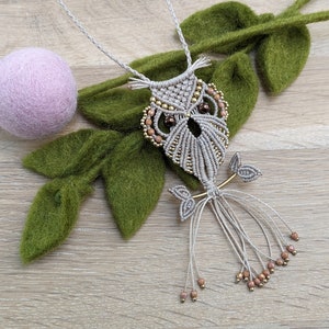 Może przedstawiać: Naszyjnik z beżowym wisiorem w kształcie sowy wykonanym z makramu z brązowymi koralikami. Naszyjnik wisi na gałęzi z zielonymi liśćmi i różową kulką z wełny.