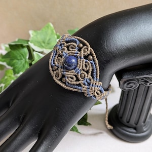 Könnte beinhalten: Ein Makramee-Armband mit einem zentralen blauen Stein und aufwendigen Webmustern in Blau und Beige. Das Armband wird auf einer schwarzen Mannequin-Hand präsentiert, die die detaillierte Handwerkskunst und das Design des Schmucks hervorhebt.