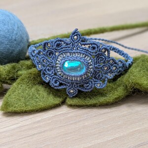 Könnte beinhalten: Ein blaues Makramee-Armband mit einem türkisfarbenen Edelstein in der Mitte. Das Armband ist mit einer Knotentechnik gefertigt und hat ein zartes, filigranes Design.