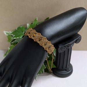 Puede incluir: Una pulsera de macramé con cordón marrón y pequeñas cuentas redondas de color ámbar. La pulsera se exhibe en un soporte de joyería negro en forma de mano. El fondo incluye vegetación y una columna decorativa negra.