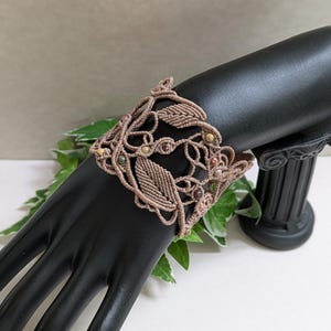Puede incluir: Un brazalete de macramé marrón claro, con un diseño de hojas y vides y pequeñas cuentas. El brazalete se exhibe en un soporte negro con forma de mano. Una pieza de joyería hecha a mano única.