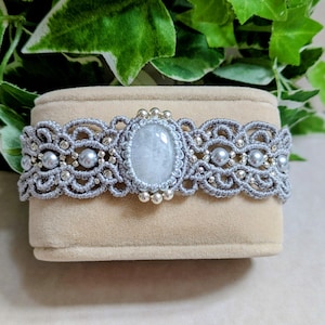 Può includere: Un braccialetto in macramè grigio con una pietra centrale ovale di colore chiaro. Il braccialetto presenta perline argentate e perle finte. Il design è intricato e dettagliato, mostrando un'estetica artigianale. Il braccialetto è esposto su una superficie beige.