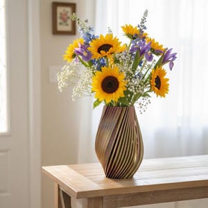 May include: A wooden vase with a unique, striped design holds a vibrant bouquet of sunflowers, irises, and other flowers. The vase sits on a wooden table. The flowers are yellow, purple, blue, and white.