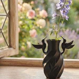 May include: A matte black vase featuring a twisted, spiral body with embossed leaf patterns and two sculpted hummingbird figures attached to the sides. The vase holds a single stem of purple delphinium flowers with small green leaves.
