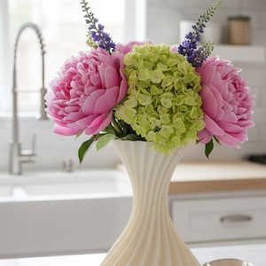 May include: A cream-colored, swirled vase holds a vibrant bouquet of pink peonies, green hydrangeas, and purple flowers. The vase sits on a white countertop in a bright kitchen setting, with a sink and window in the background.