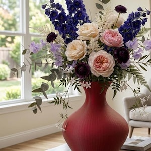 Peut inclure: Un grand vase rouge mat avec une base large et arrondie et un col étroit, rempli d'un arrangement floral vibrant. Le bouquet comprend des delphiniums bleu foncé, des roses de jardin rose pâle et crème, des renoncules violet foncé et de l'eucalyptus.